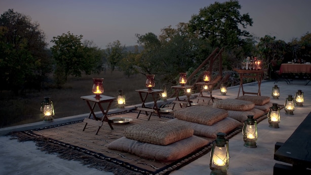 An outdoor seating area with tables and cushions, surrounded by lanterns and trees.