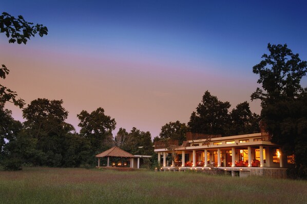 A large, modern house with a spacious porch and multiple rooms, surrounded by a grassy field and trees. - Mahua Kothi, Bandhavgarh National Park (Manpur)