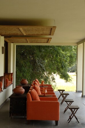 A covered outdoor seating area with orange sofas and wooden tables, surrounded by greenery and a view of a body of water. - Mahua Kothi, Bandhavgarh National Park (Manpur)