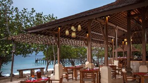 An outdoor dining area with wooden tables and chairs, a thatched roof, and a view of the ocean. - Nusabay Menjangan by WHM (Sumberklampok)