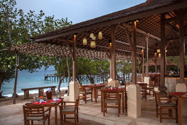 An outdoor dining area with wooden tables and chairs, a thatched roof, and a view of the ocean. - Nusabay Menjangan by WHM (Sumberklampok)