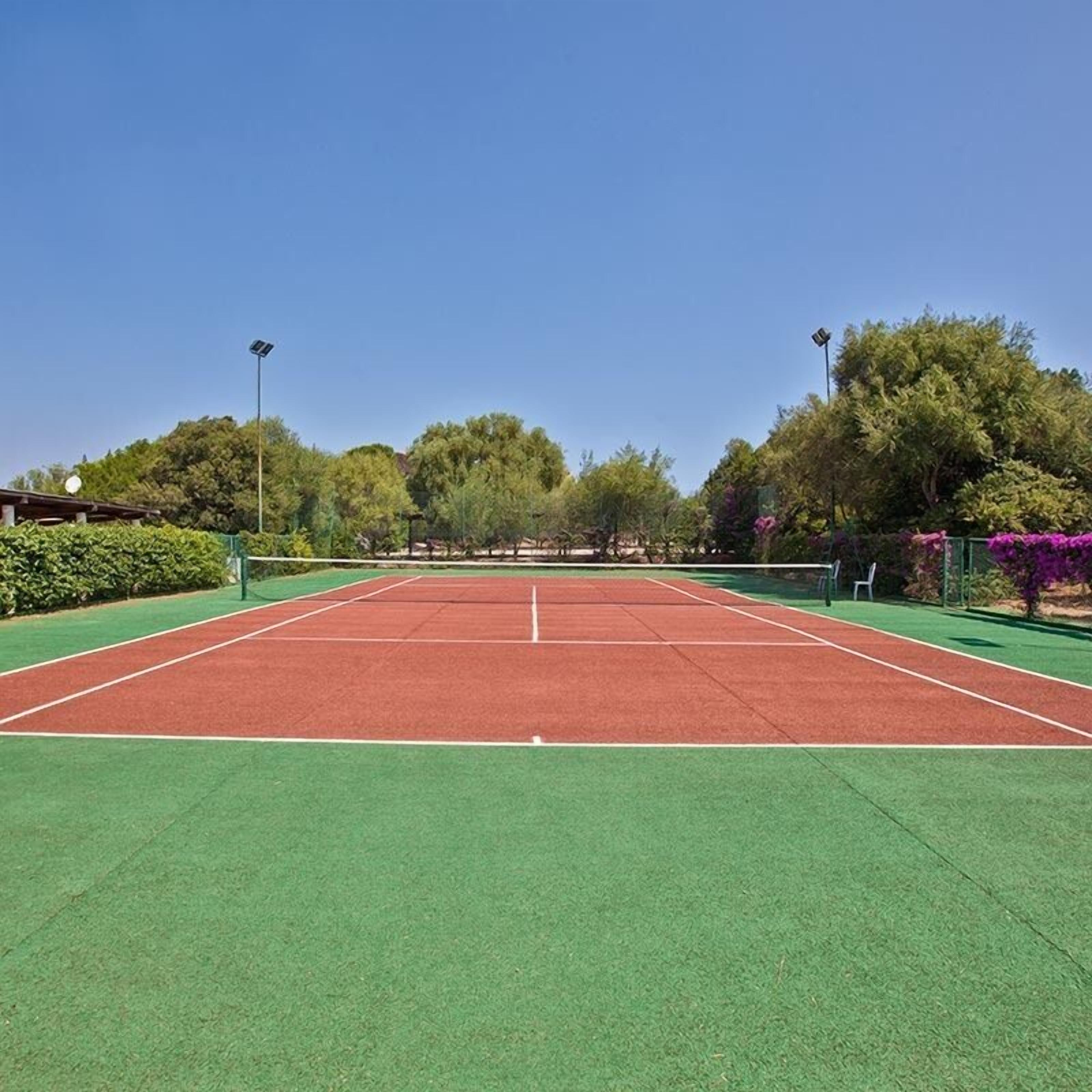 A tennis court with green and red surfaces, surrounded by trees and shrubs.