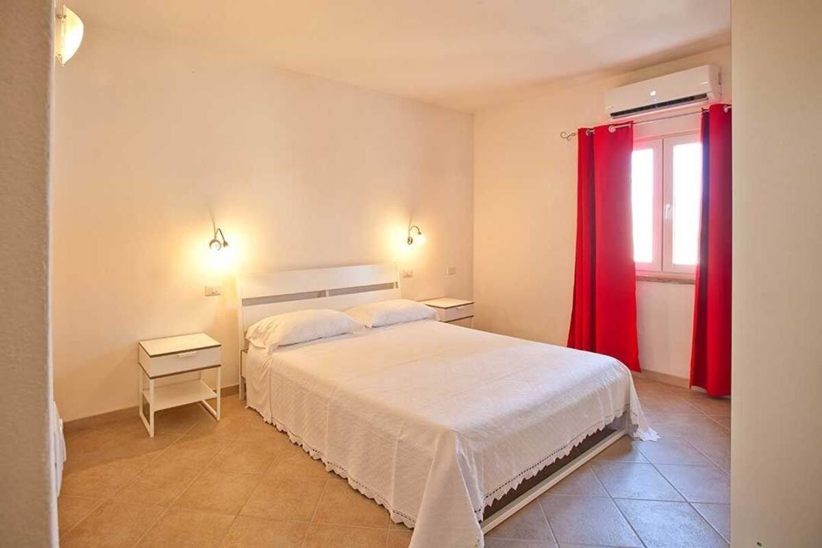 A bedroom with a bed, two bedside tables, a nightstand, a window with red curtains, and an air conditioning unit.