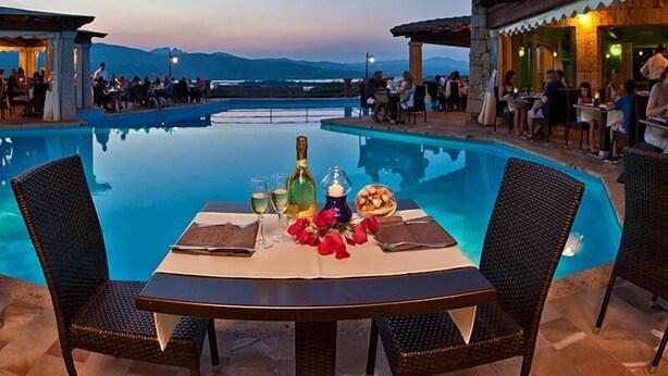 Un área de comedor junto a la piscina con una mesa preparada para dos, rodeada de sillas de mimbre, con vistas a una piscina y montañas a la distancia.
