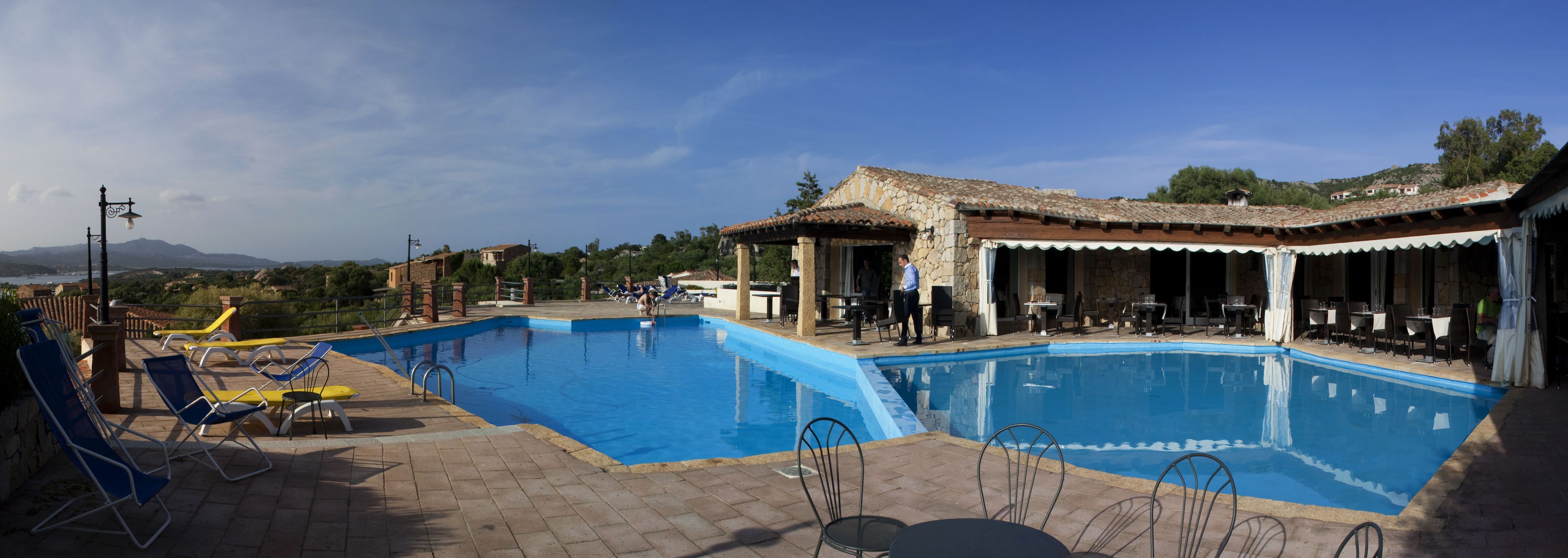 Una piscina con sillas de descanso y mesas, rodeada de edificios de piedra y un cielo despejado.