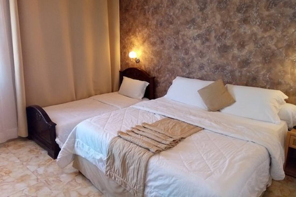 Cama doble con sábanas blancas y una manta color beige, cabecera de madera, mesita de noche y una lámpara de pared.