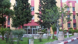 Un edificio de varios pisos con fachada roja y amarilla, una fuente y un jardín bien cuidado con diversas plantas y un sendero de piedra.