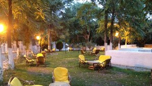 Un espacio al aire libre con sillas y mesas de mimbre, rodeado de árboles y una pared blanca.