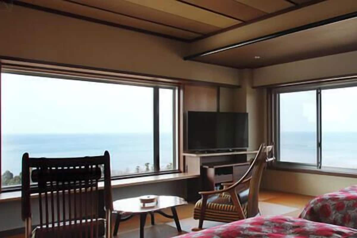 Une chambre avec un lit, une télévision, une chaise, une table et de grandes fenêtres donnant sur l’océan.