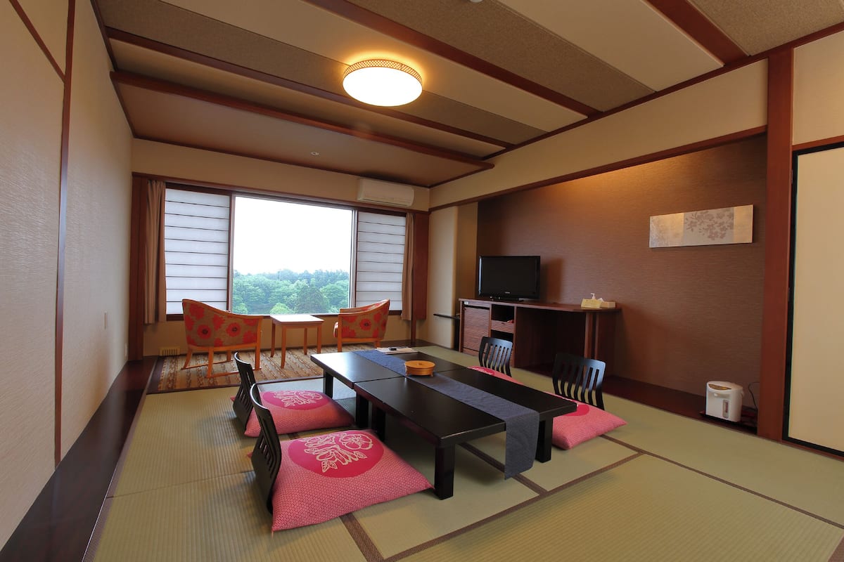 Une pièce de style japonais traditionnel, avec un sol recouvert de tatamis, une table basse, des chaises, un téléviseur et une vue par la fenêtre.