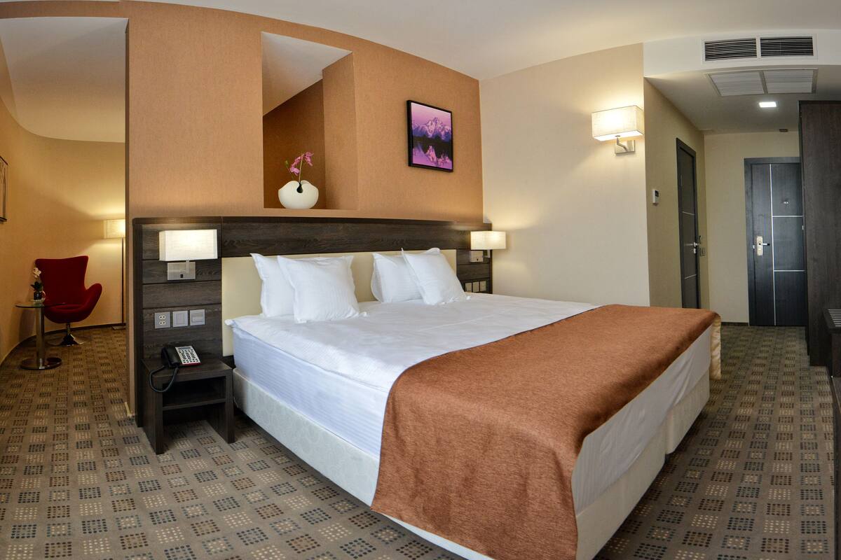 Une chambre d’hôtel avec un grand lit, une table de chevet avec un téléphone, une chaise rouge, un tableau accroché au mur et une porte donnant sur la salle de bain.