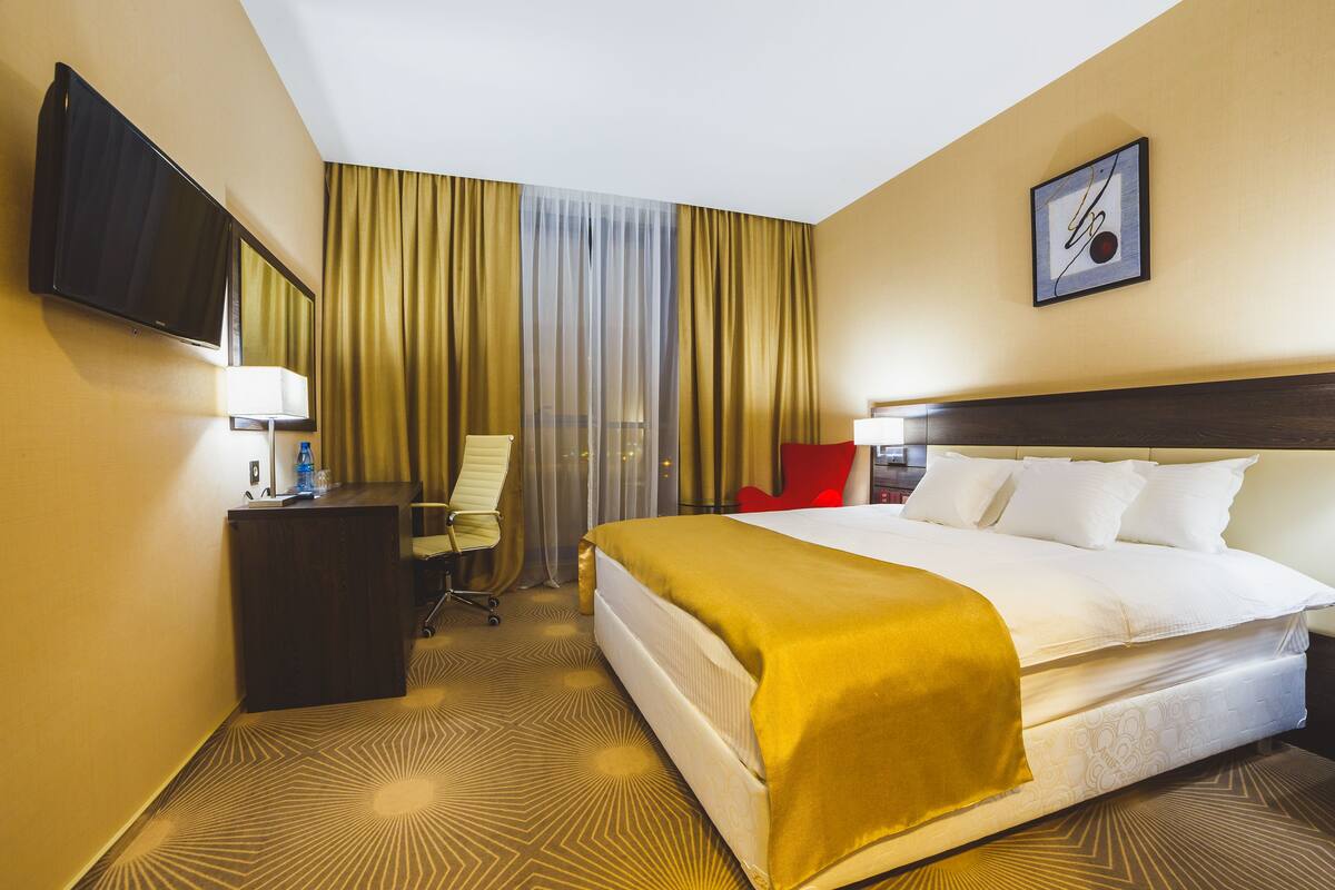 Une chambre d’hôtel avec un lit, un bureau, une chaise, une télévision et une fenêtre avec des rideaux.