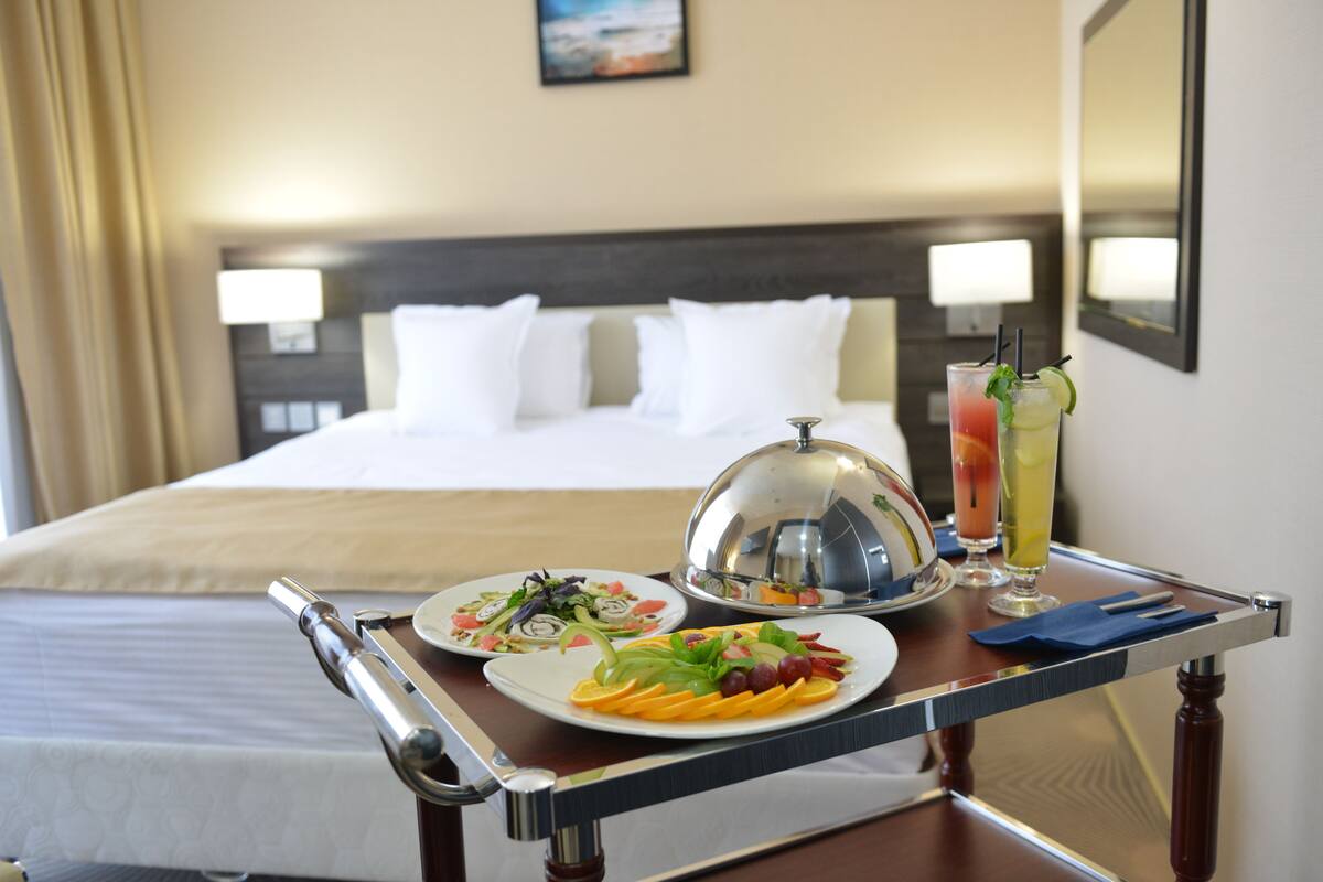 Une chambre d’hôtel avec un lit soigneusement fait, un chariot de service avec des assiettes remplies de nourriture et des boissons, et une table de chevet avec une lampe.