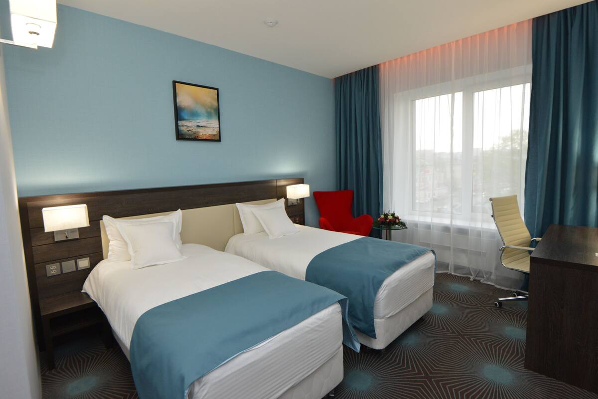Une chambre d’hôtel avec deux lits, un bureau, une chaise, une table de chevet avec des lampes et un tableau au mur.