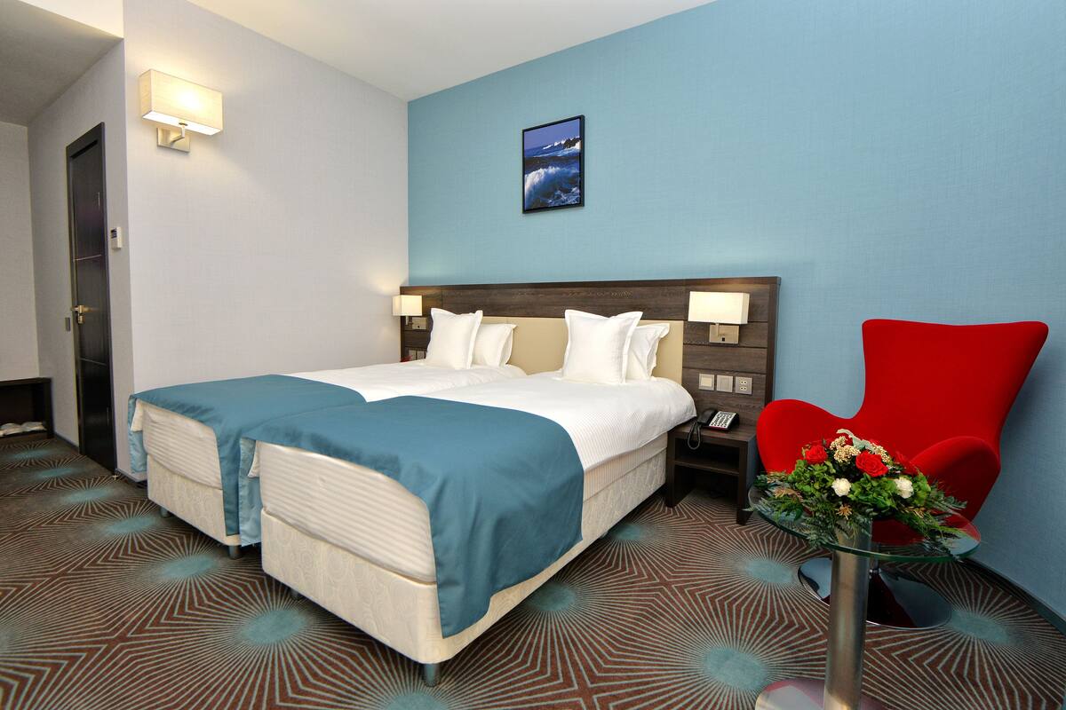 Une chambre d’hôtel avec un lit, une chaise rouge, une table de chevet et une lampe fixée au mur.