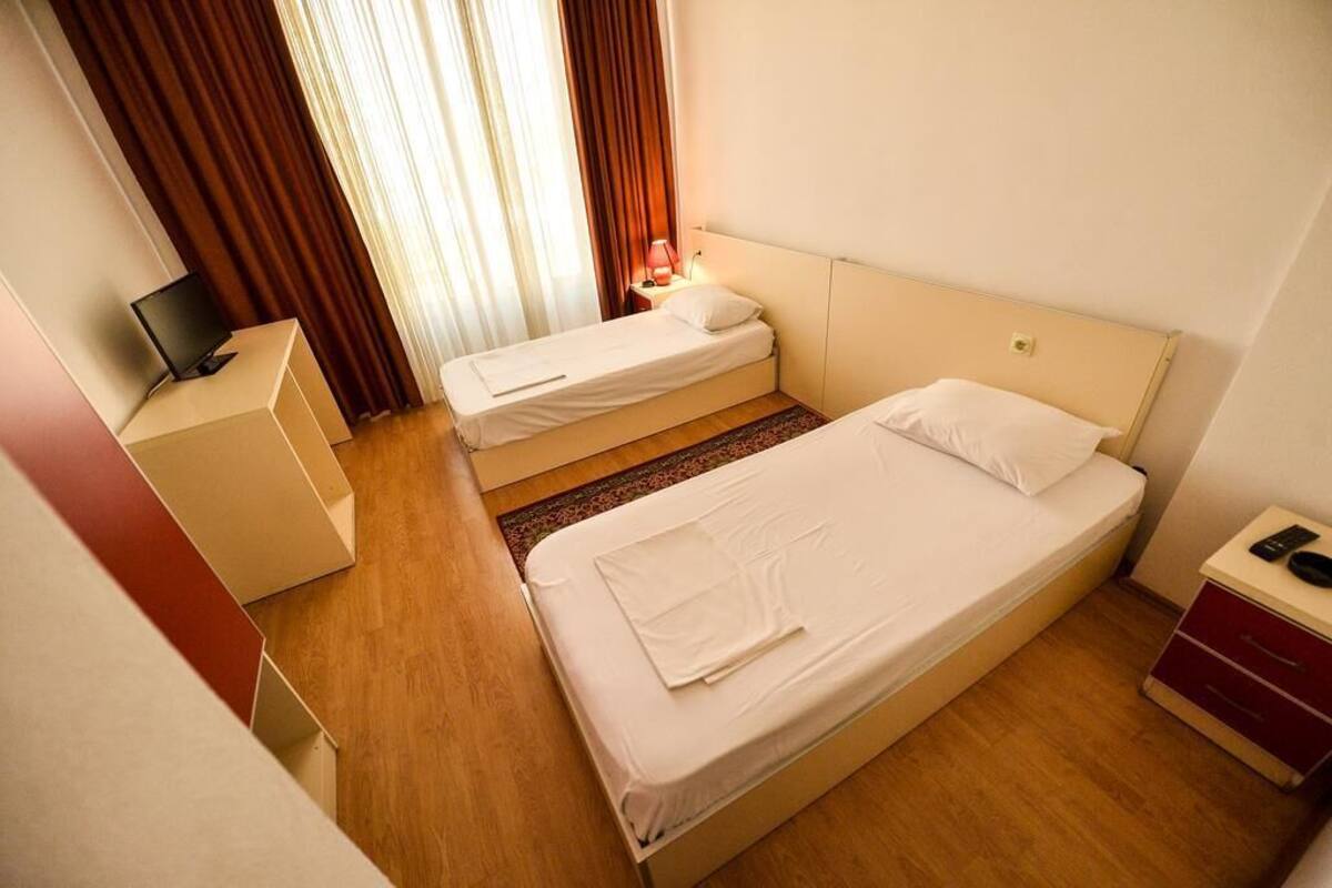 Quarto de hotel com duas camas de solteiro, uma escrivaninha com computador e uma mesa de cabeceira com abajur.