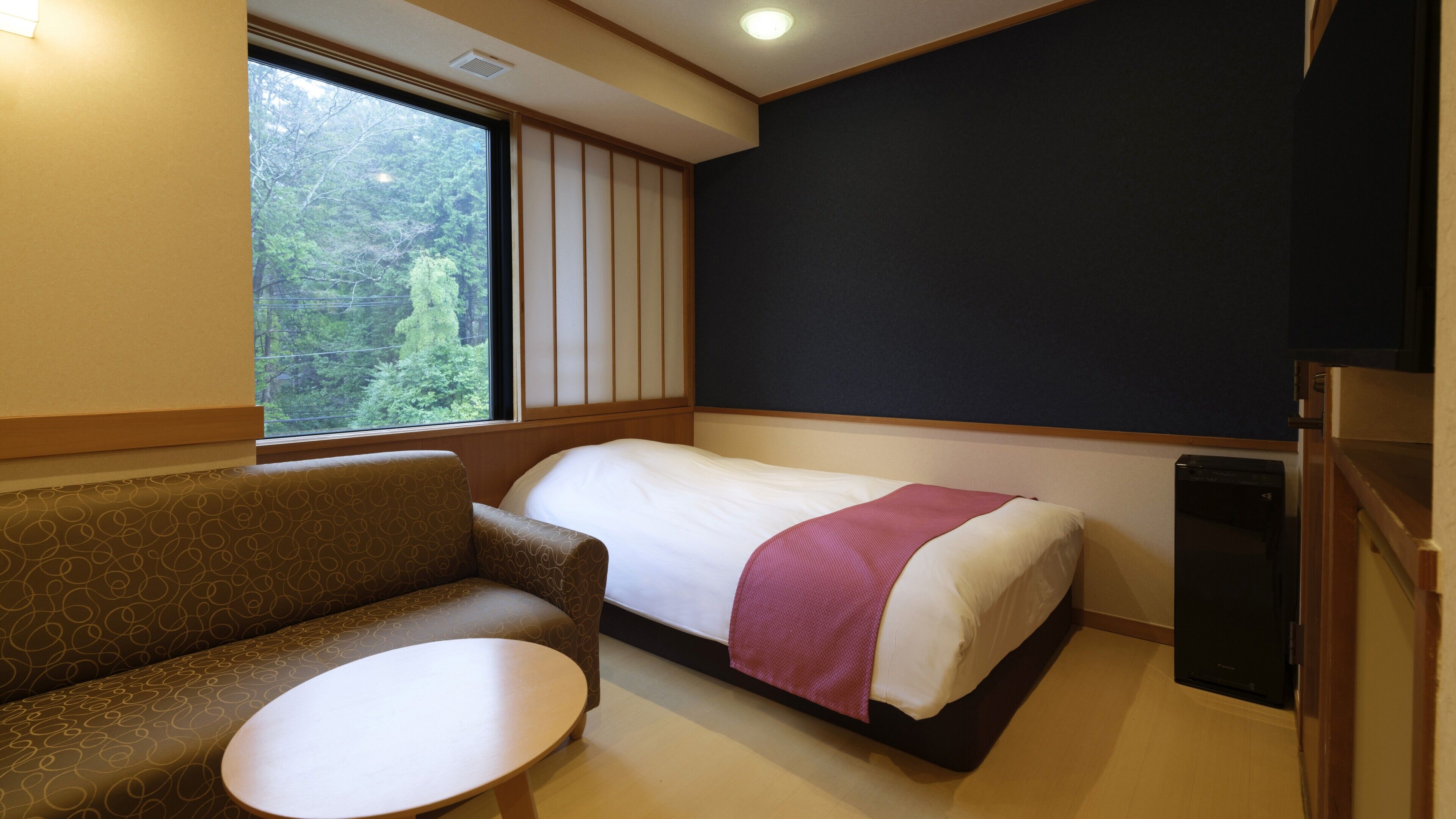 A single bed with a white and pink bedspread, a small round table, and a sofa in a room with a large window and a dark wall.