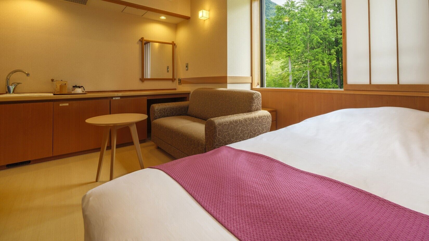 A hotel room with a bed, a chair, a small table, a sink, and a window with a view of trees.