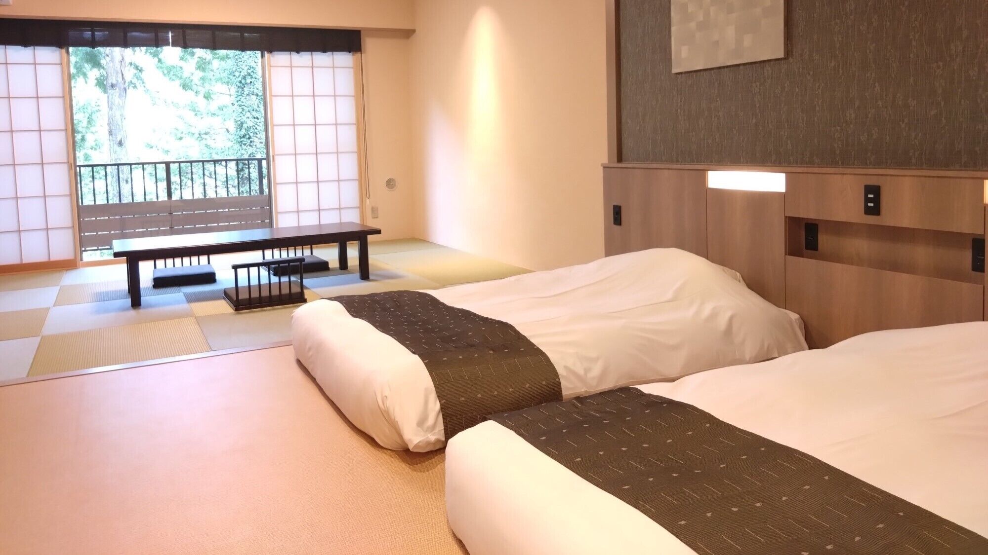 Une chambre d’hôtel avec un sol recouvert de tatamis, deux lits, un banc et une porte coulissante donnant sur un paysage verdoyant.