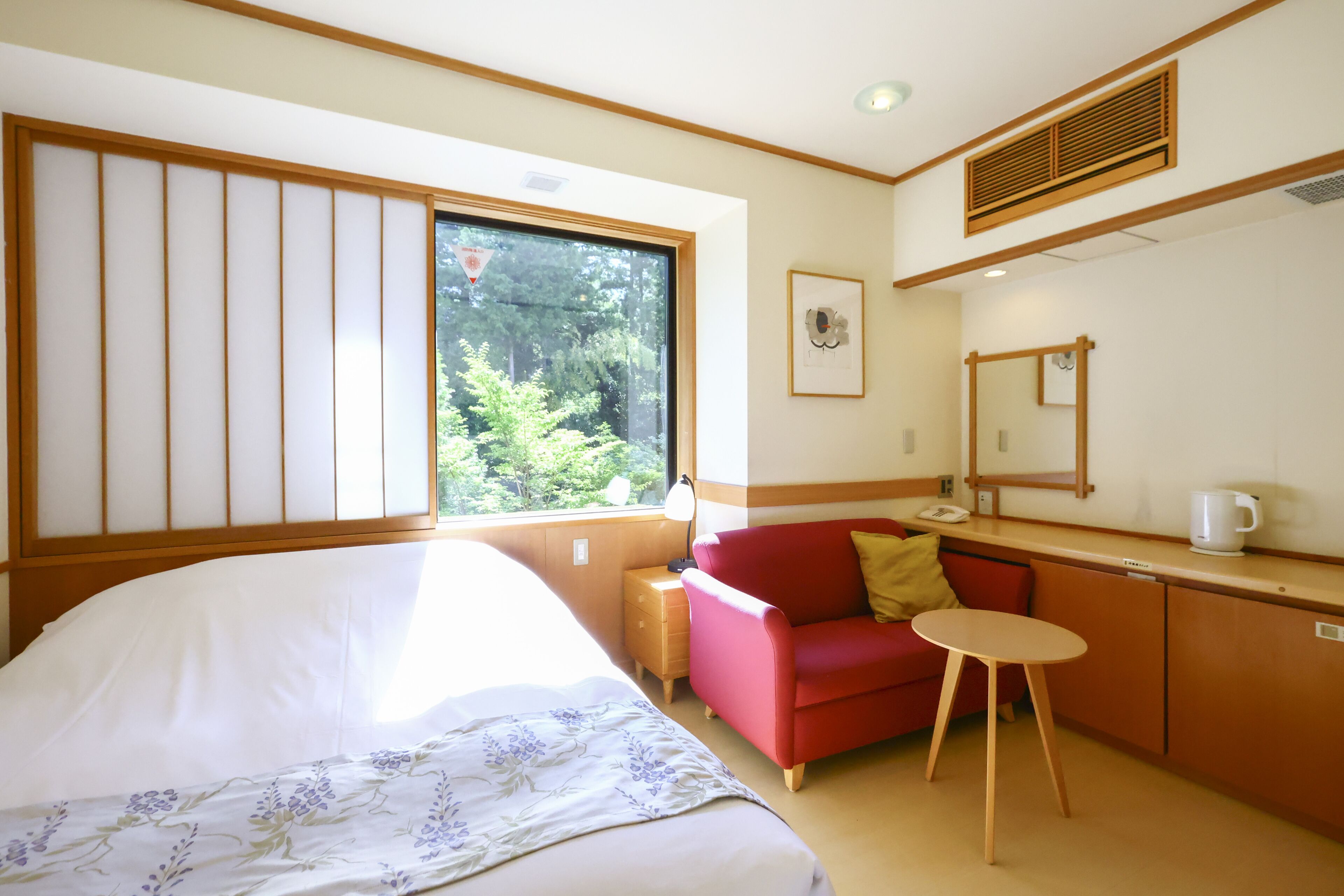Een hotelkamer met een bed, een rode bank, een klein tafeltje, een spiegel en een raam met uitzicht op bomen.