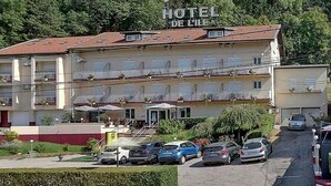 A hotel with a sign reading "HOTEL DE L'ILE". It features a two-story building with balconies, a parking area with several cars, and a garden area with plants and a pathway. - Hôtel de l'île (Custines)