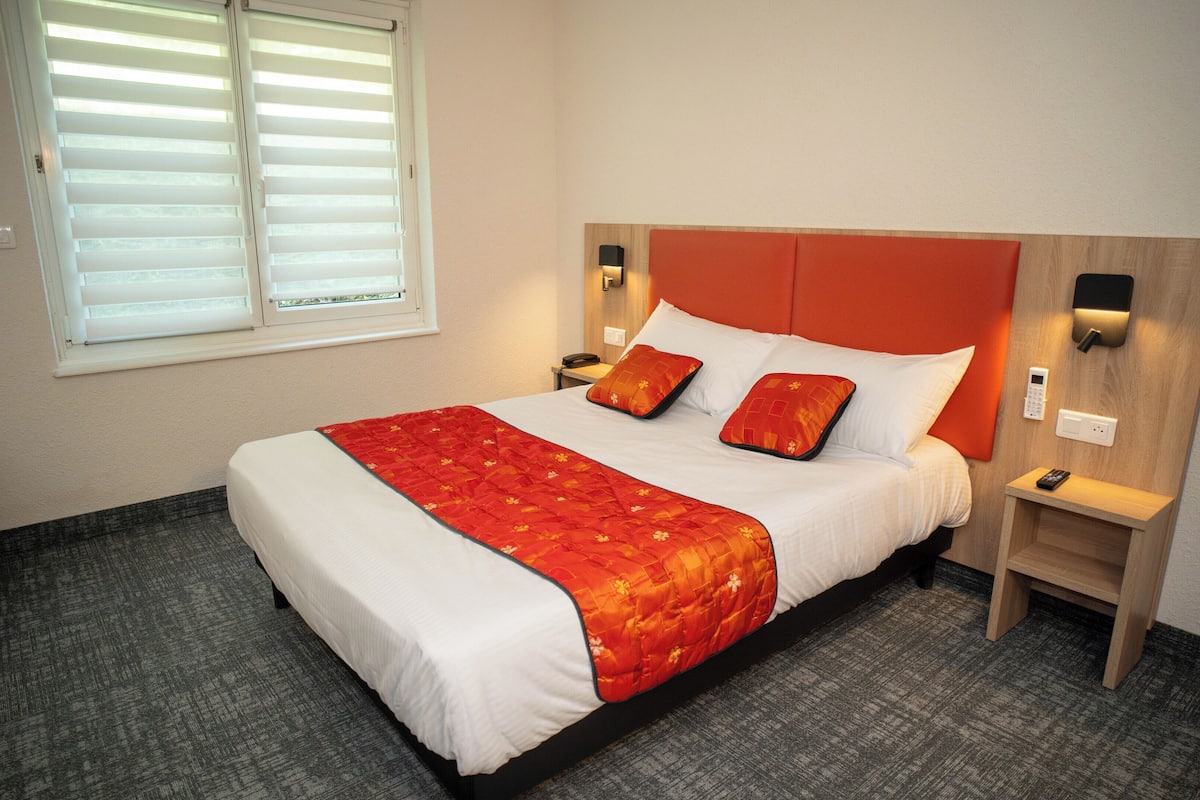 A neatly made bed with a red and white bedspread, two pillows, and a wooden headboard. There is a nightstand with a phone and a lamp, and a window with blinds.