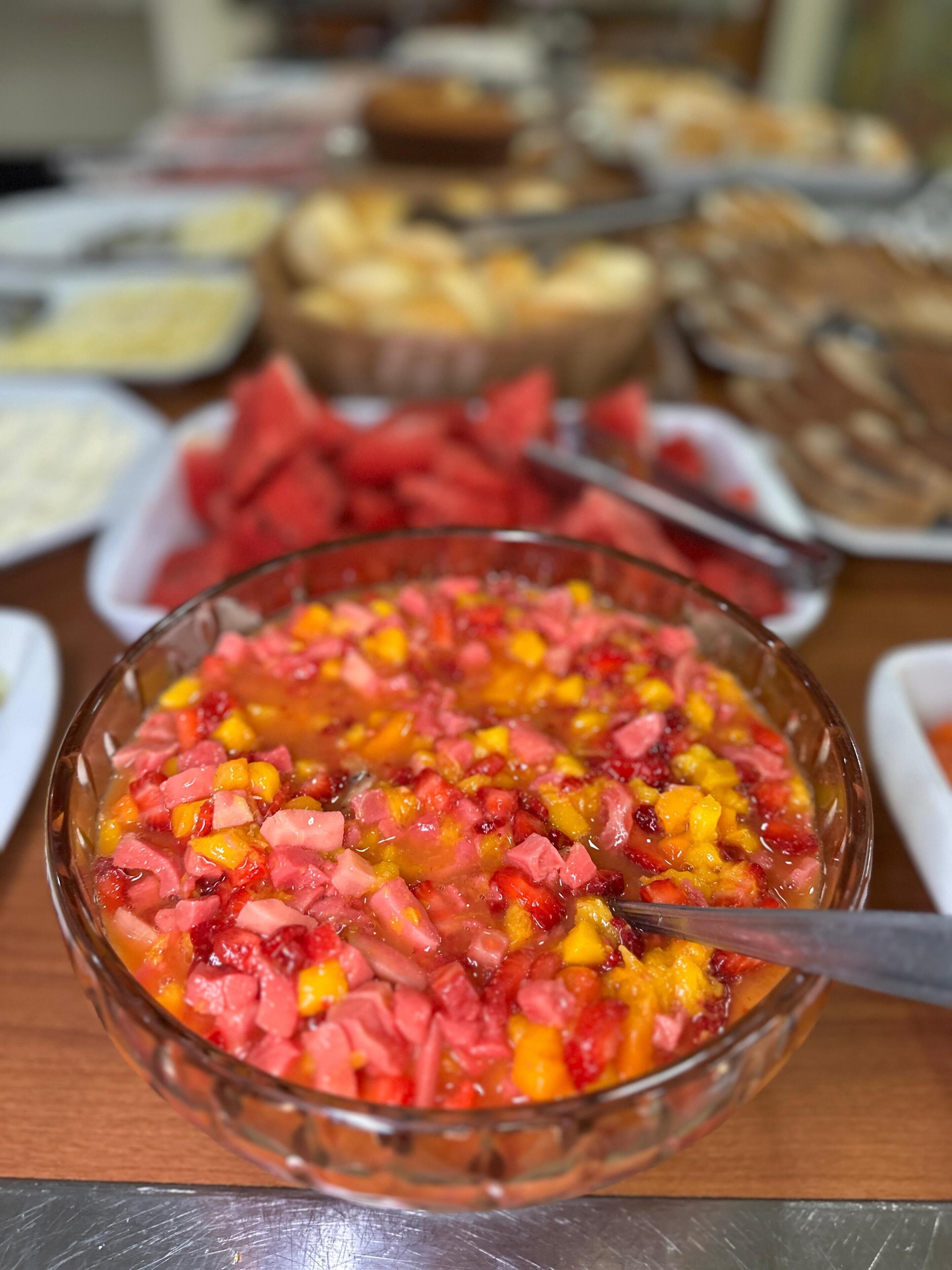 Un bol de vidrio lleno de ensalada de frutas y carne, rodeado de varios platos y utensilios sobre una mesa.