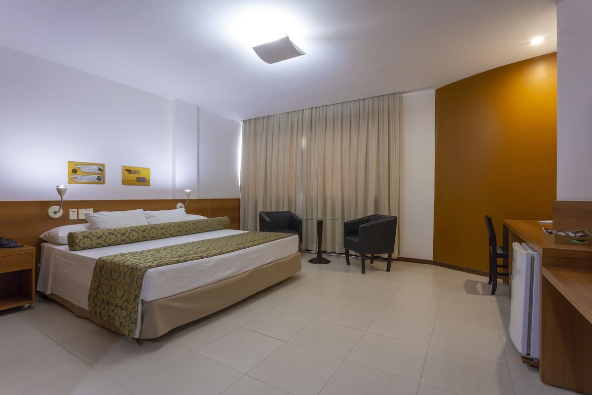 Una habitación de hotel con cama, dos sillas, una mesita, un escritorio y un refrigerador.