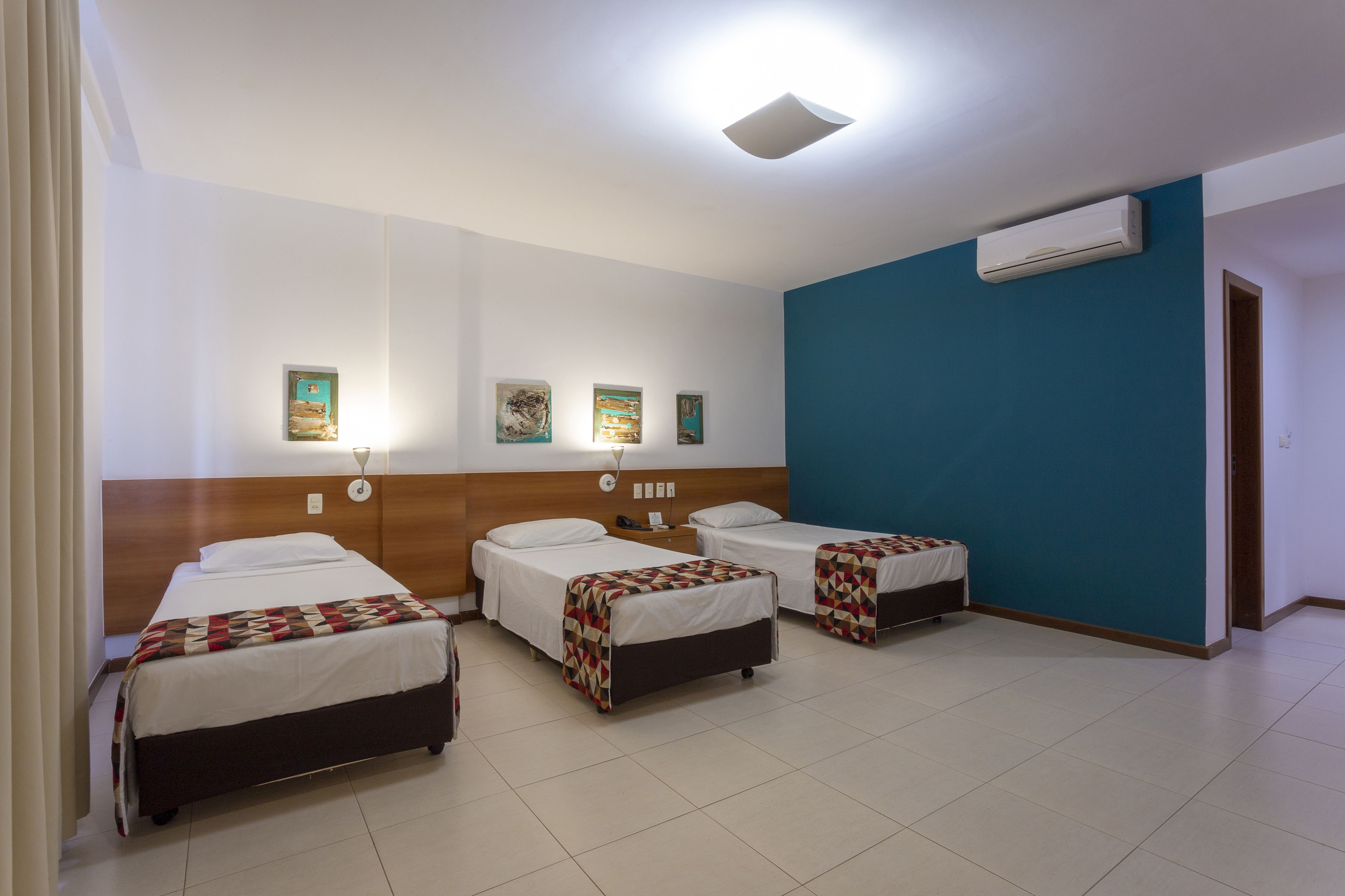 Habitación de hotel con tres camas individuales, cabecera de madera, pared de acento azul y un equipo de aire acondicionado.