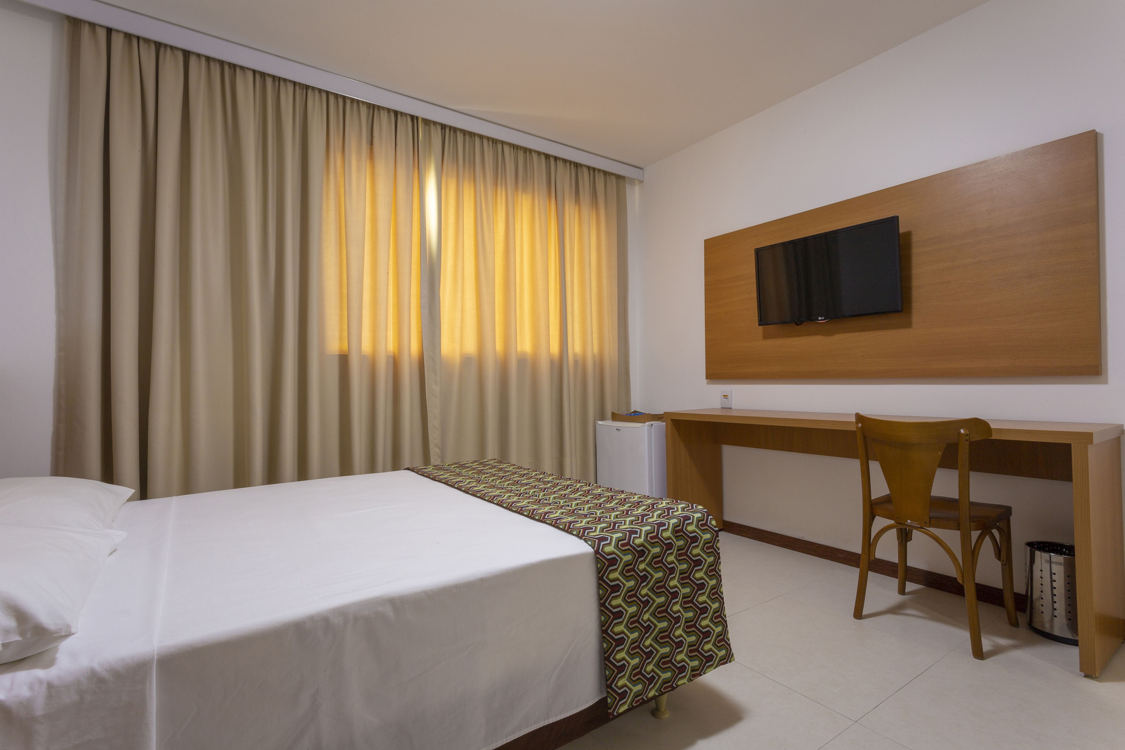 Habitación de hotel con cama, escritorio, silla, televisor y ventana con cortinas.