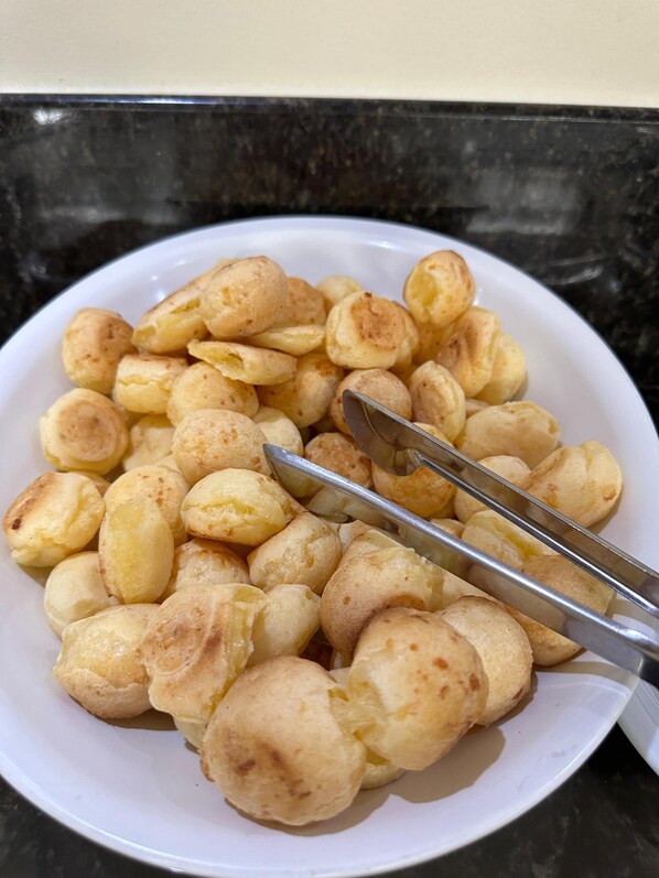 A white bowl filled with small, round, golden-brown pastries, likely cheese puffs, with metal tongs resting on top. - Hotel Serra Grande - Serra/ES (Serra)