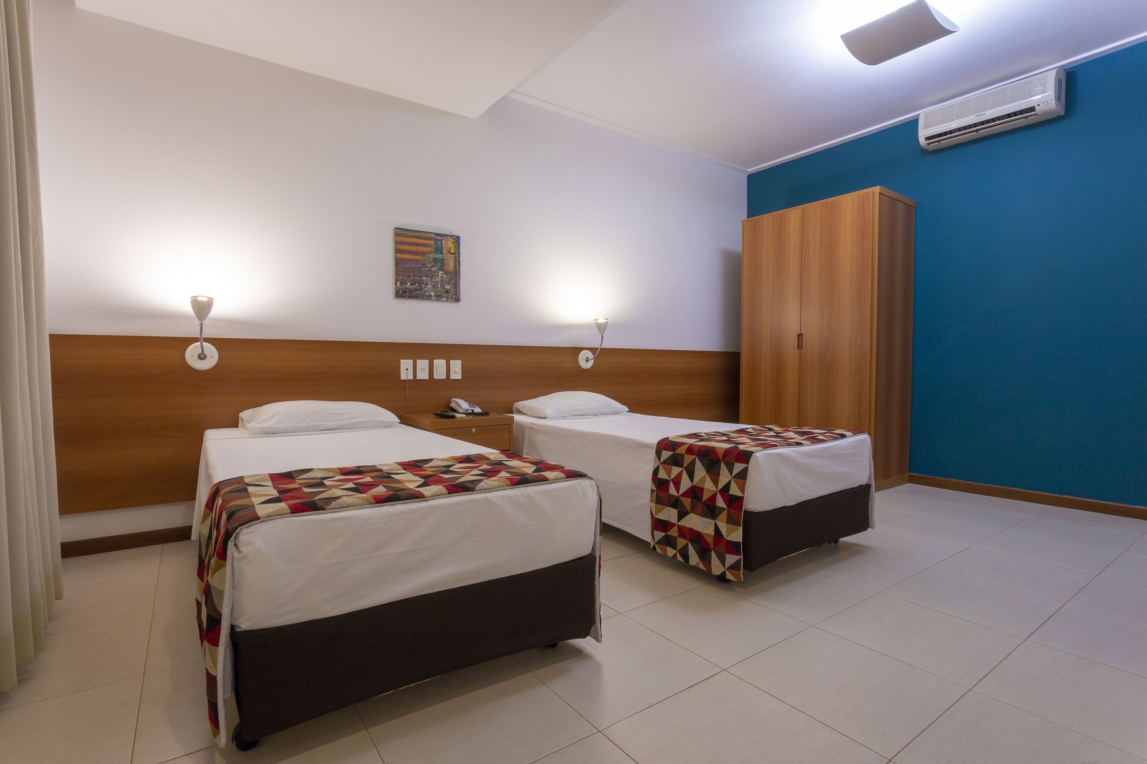 A hotel room with two beds, a wooden wardrobe, and a cityscape painting on the wall.