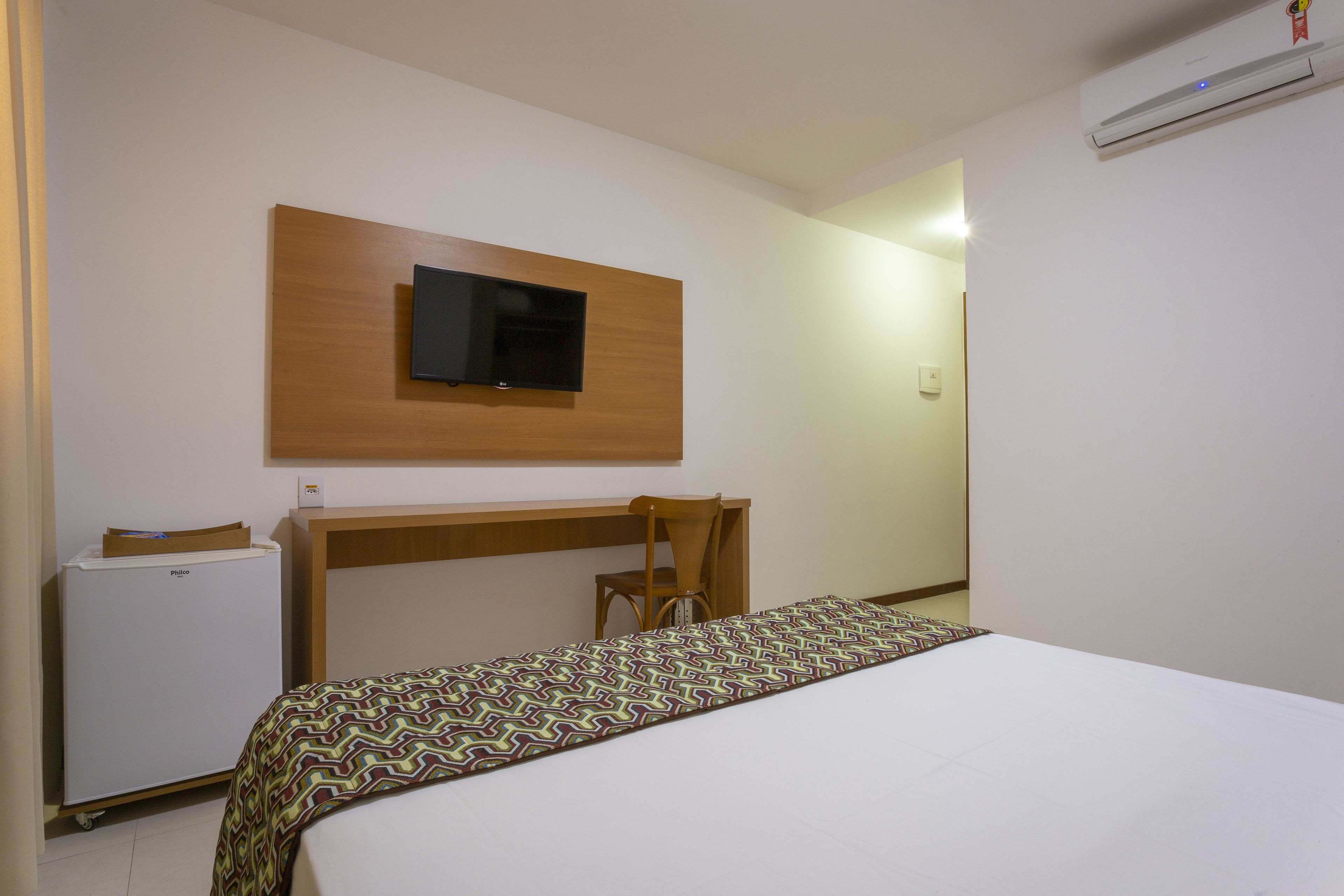 A hotel room with a bed, a desk, a chair, a television mounted on the wall, and an air conditioning unit.