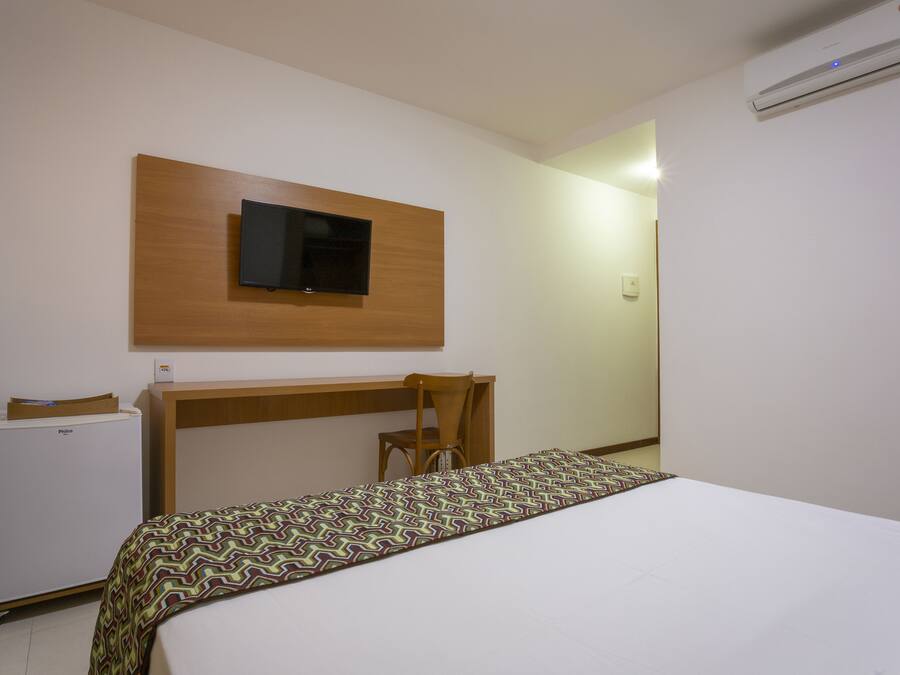 Quarto de hotel com cama, escrivaninha, cadeira, televisão fixada na parede e ar condicionado.
