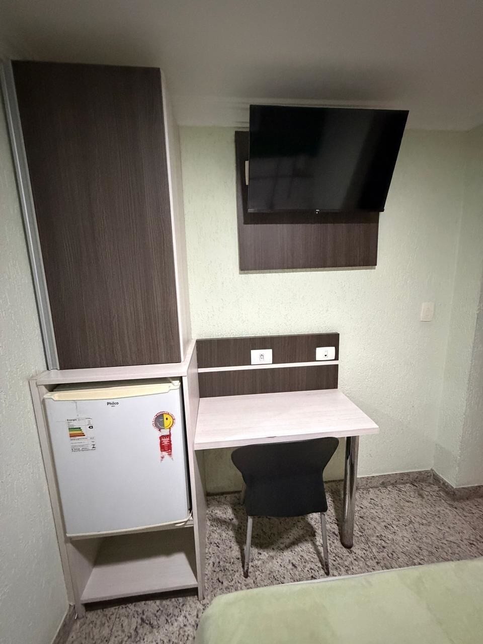 Una habitación pequeña con escritorio, silla, televisor montado en la pared y refrigerador.