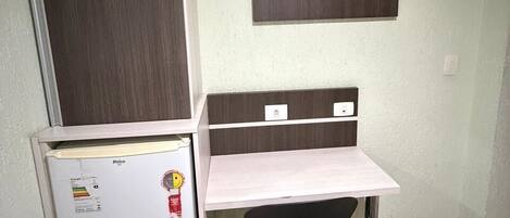 Una habitación pequeña con escritorio, silla, televisor montado en la pared y refrigerador.