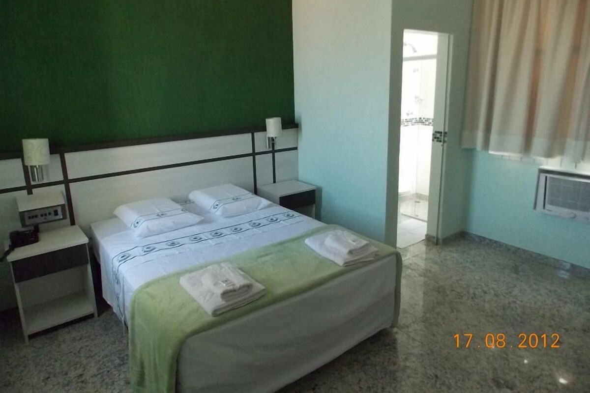Une chambre d’hôtel comprenant un lit, des tables de chevet, une commode, un mur vert, une fenêtre avec des rideaux et un appareil de climatisation.