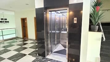 Um saguão com piso xadrez, um elevador e uma planta em vaso.