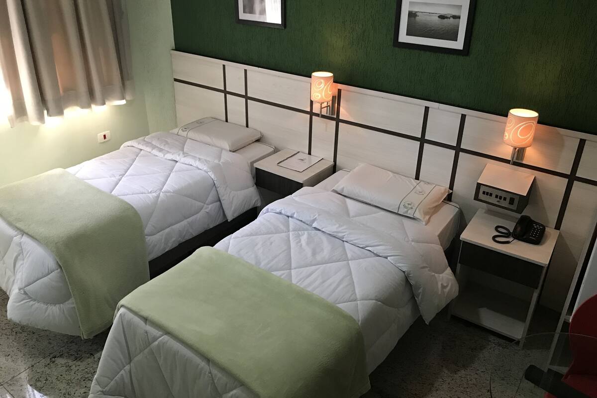 Ein Hotelzimmer mit zwei Betten, jeweils mit Kopfkissen und Decke. An den Nachttischen stehen Lampen und ein Telefon. Die Wände sind grün, und es sind Bilder an der Wand zu sehen.