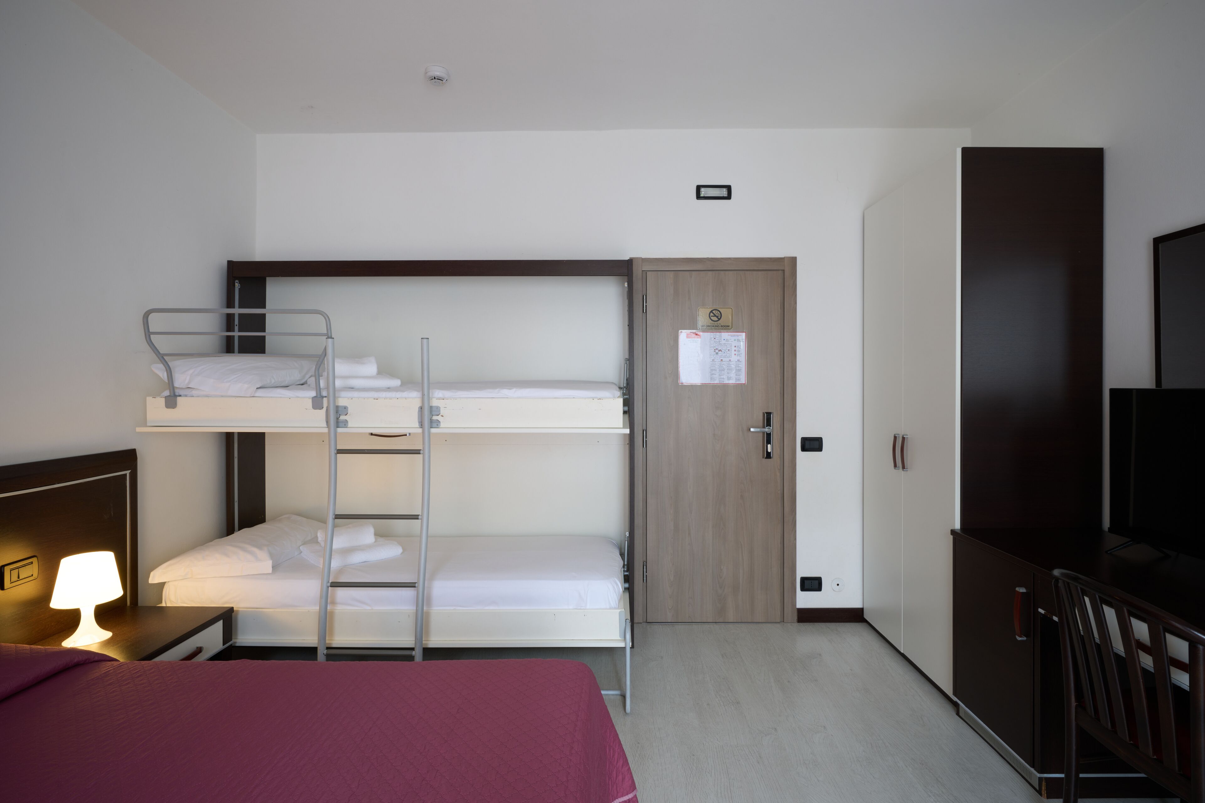 A hotel room with bunk beds, a desk with a lamp, a TV, and a wardrobe.
