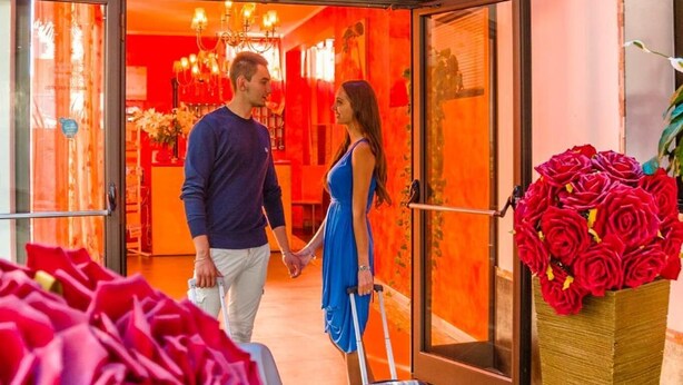 Vestíbulo de hotel con un techo naranja brillante, un jarrón grande de rosas rojas y una persona arrastrando una maleta.