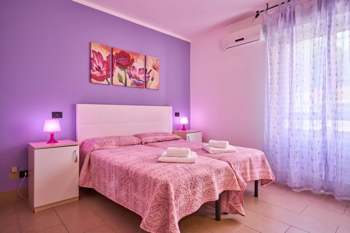 Un dormitorio con dos camas, una mesita de noche con lámpara, otra mesita de noche con lámpara y una ventana con cortinas translúcidas.