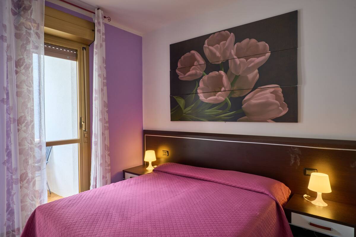 Una cama bien hecha con colcha rosa, un cuadro de flores rosadas colgado en la pared y dos lámparas de noche sobre una mesita.