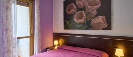 A neatly made bed with a pink bedspread, a wall-mounted painting of pink flowers, and two bedside lamps on a nightstand.