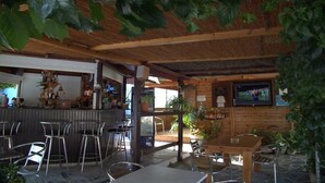 An outdoor cafe with wooden tables and metal chairs, a bar area with shelves of bottles, and a TV mounted on the wall. - Hotel Sofia (Samos)