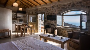 A cozy room with a stone wall, wooden ceiling beams, a large bed, a dining table, and a view through a window. - Thalori (Gortyna)