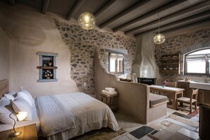 A bedroom with a stone wall, a bed, a bedside table with a lamp, a small window, and a fireplace. - Thalori (Gortyna)