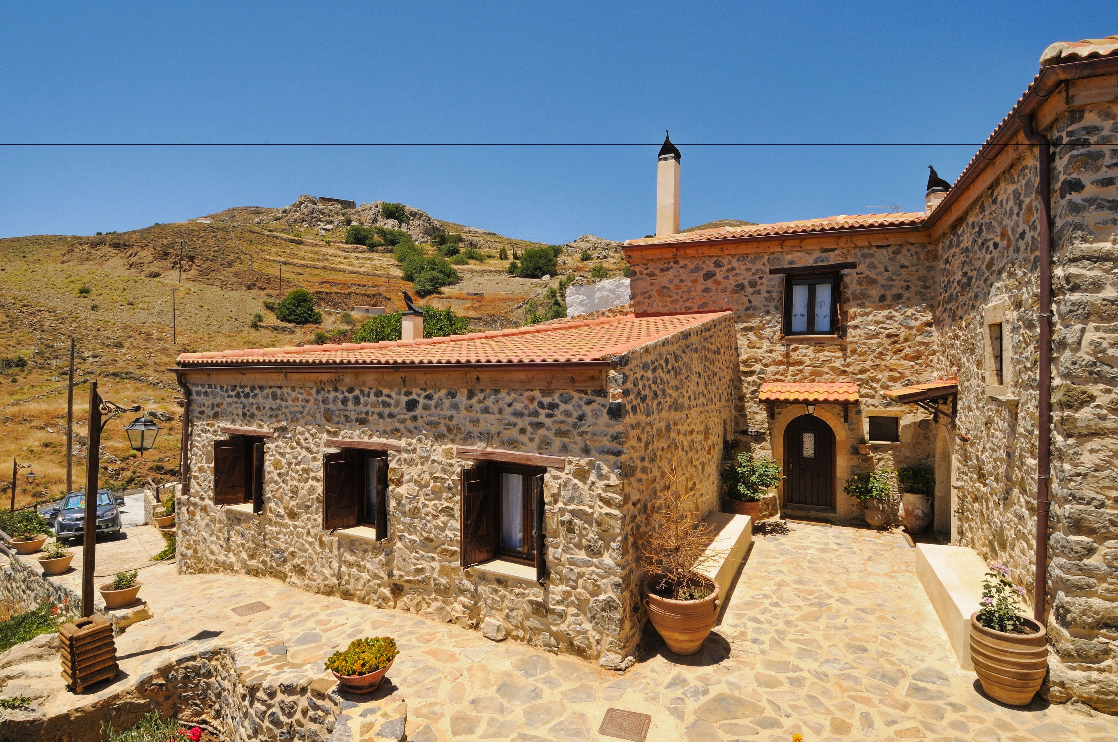 A stone building with a tiled roof, a chimney, and a small window. There are potted plants and a car parked outside.