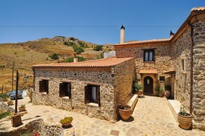 A stone building with a tiled roof, a chimney, and a small window. There are potted plants and a car parked outside. - Thalori (Gortyna)