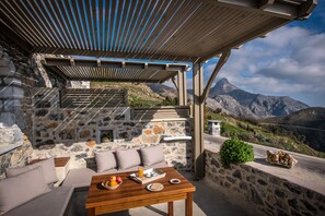 A stone-walled outdoor seating area with a wooden table and cushions, overlooking a mountainous landscape. - Thalori (Gortyna)